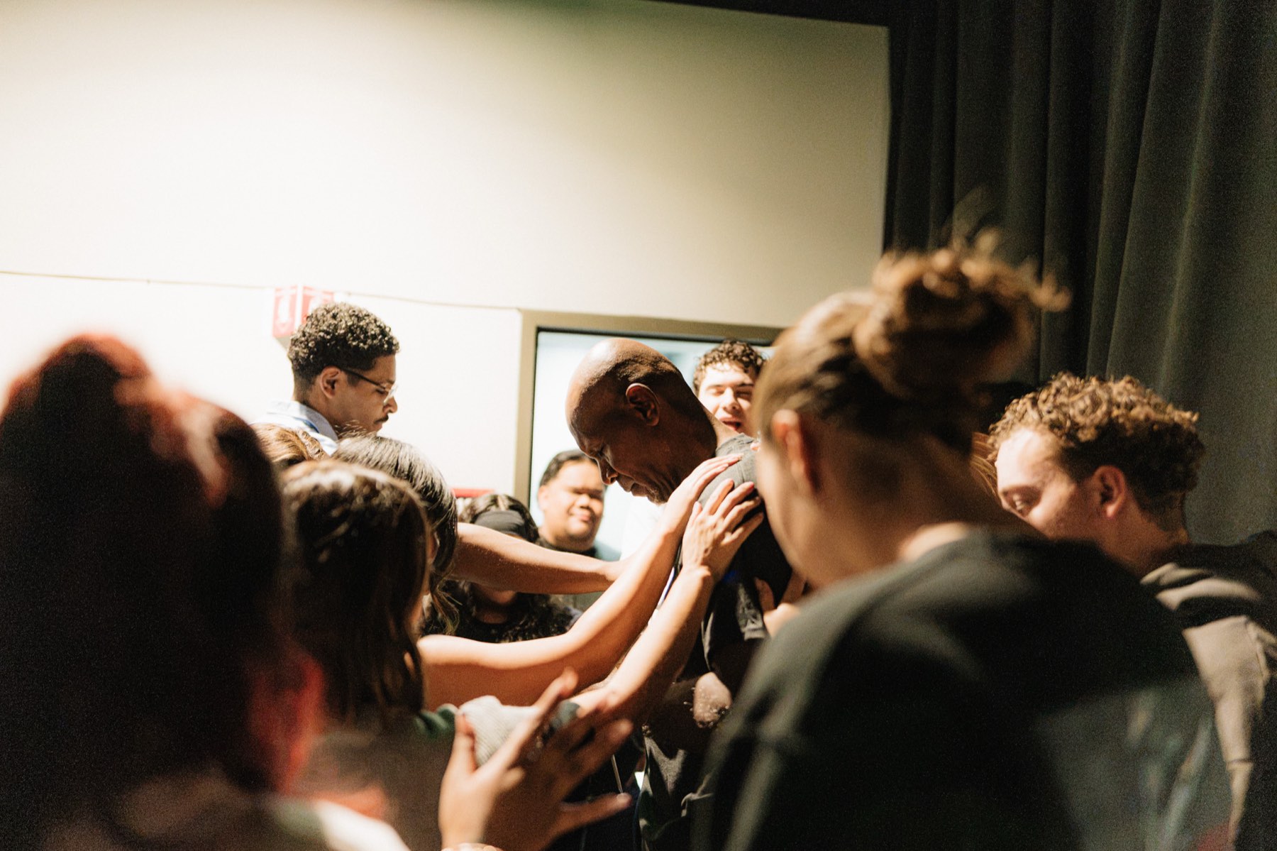 A group gathered close, praying with hands on a man's shoulders